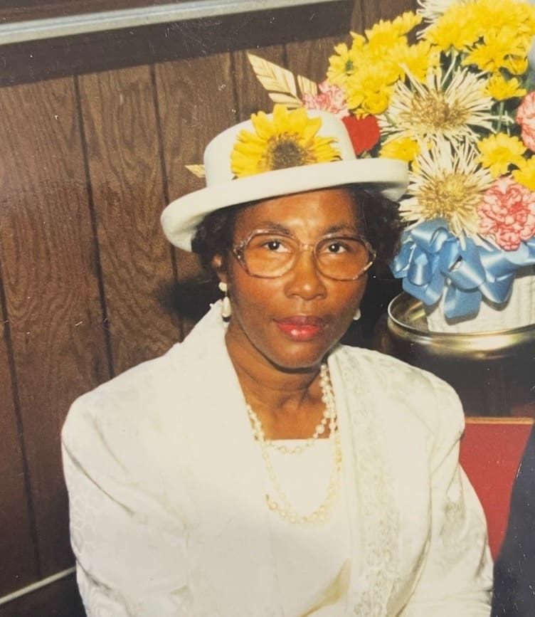 A woman wearing a hat and glasses sitting in front of flowers.